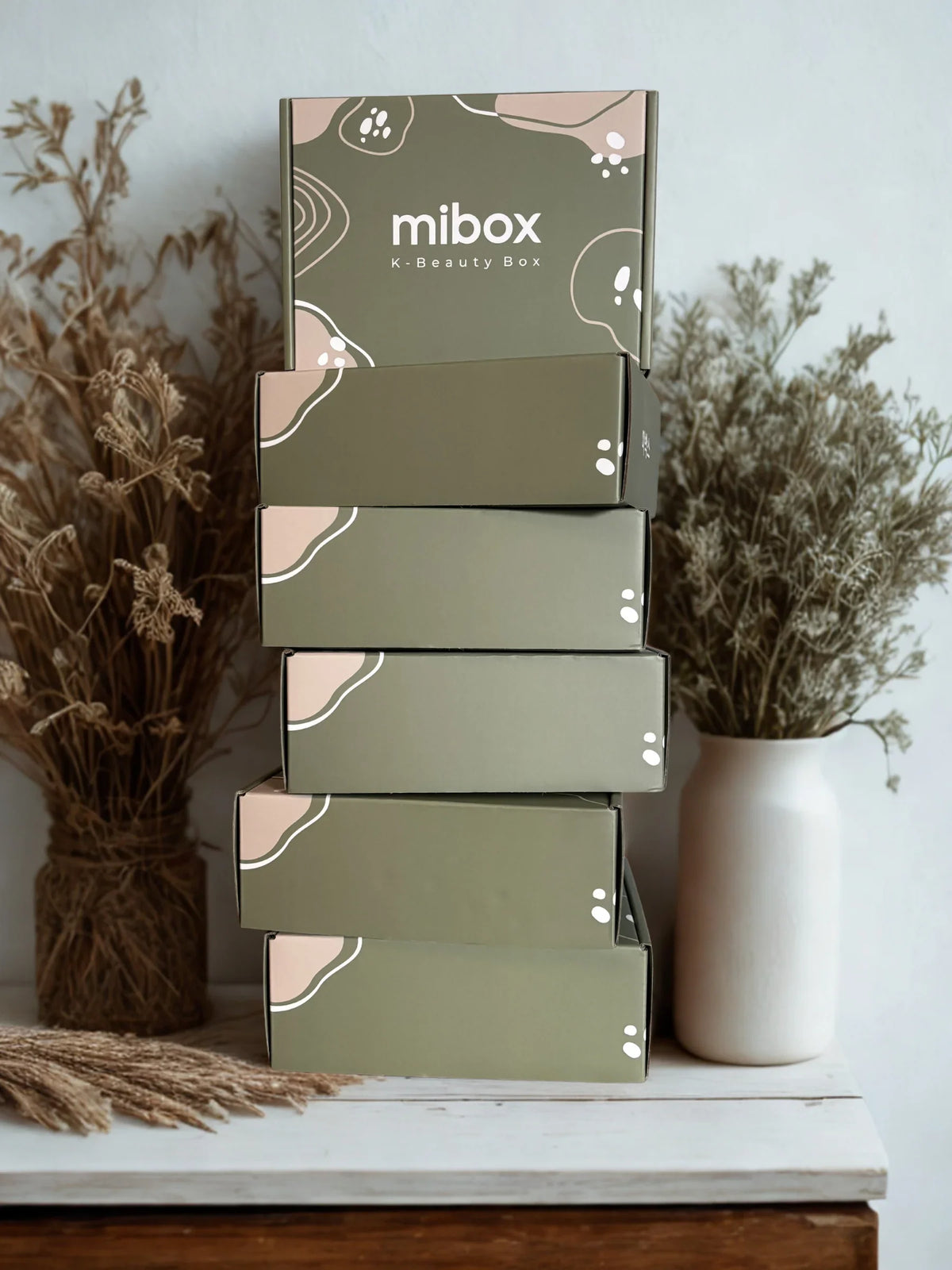 Кутии за корейска козметика Mibox, подредени върху маса, със сухи растения на заден план